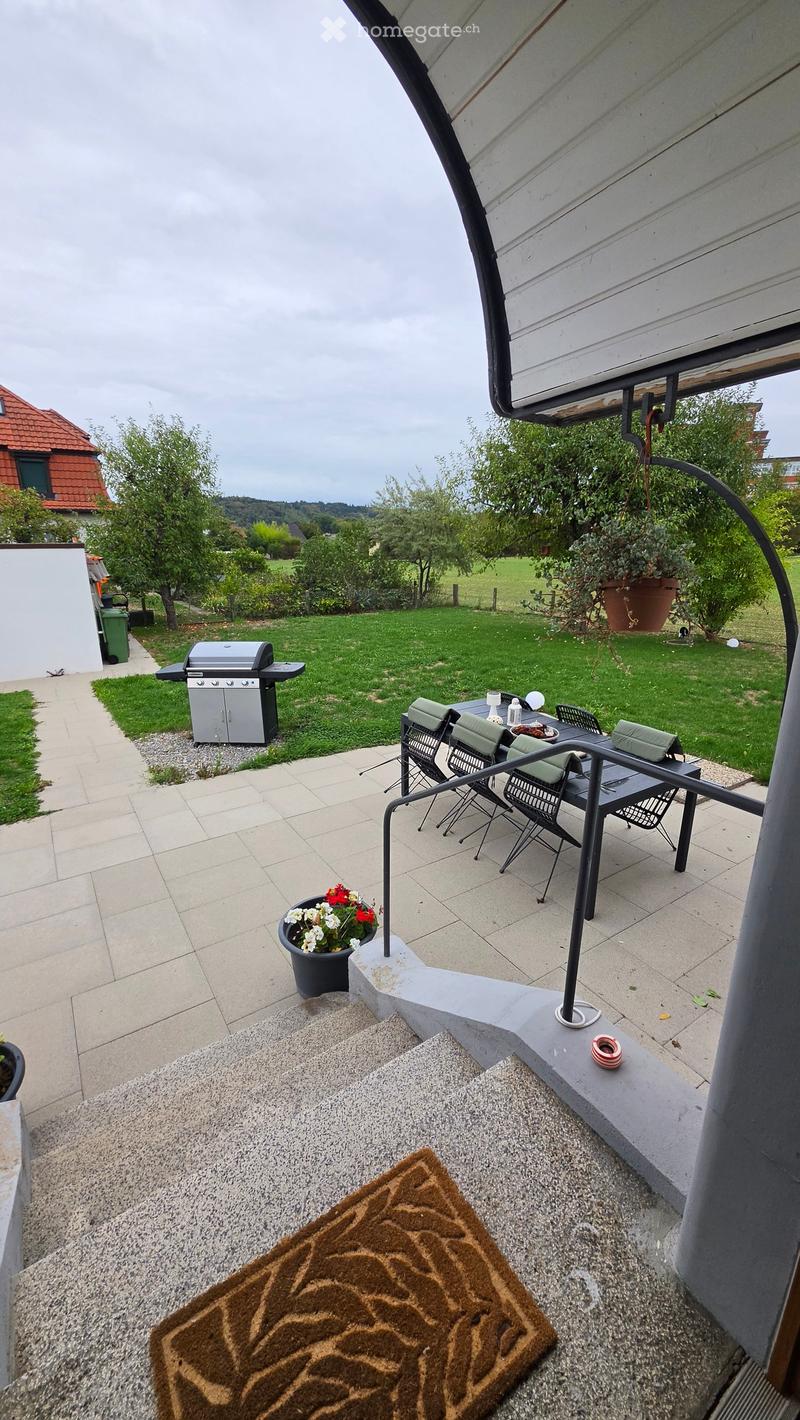 Balcony with tiled floor, metal railing, outdoor table and chairs, grill, potted plants, and a view of a green lawn and trees.