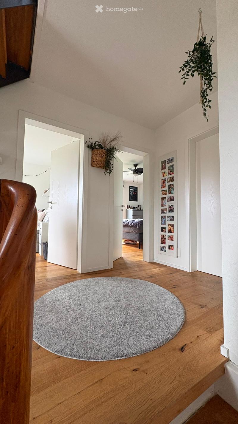 A well-lit hallway with wooden flooring, a round grey rug, and several doors leading to different rooms. There are hanging plants and a collage of photos on the wall.