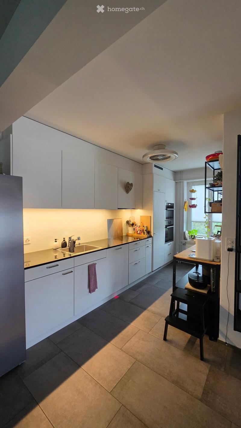 Modern kitchen with white cabinets, stainless steel sink, built-in oven, and a small table with a stool. The space features a tiled floor and ample natural light from a window.