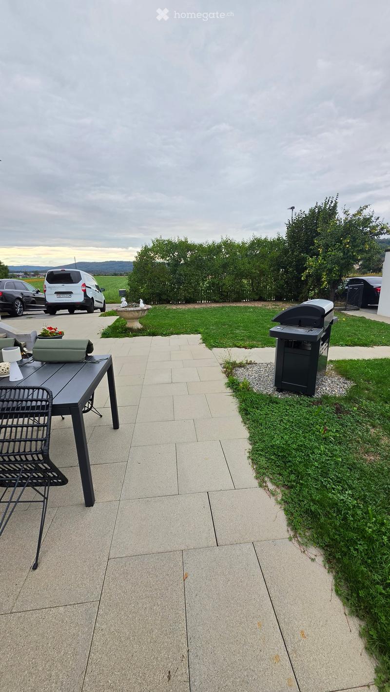 Outdoor patio area with a tiled floor, a black table and chair set, a grill, a small fountain, and a flower pot. The area is bordered by a grassy lawn and trees.