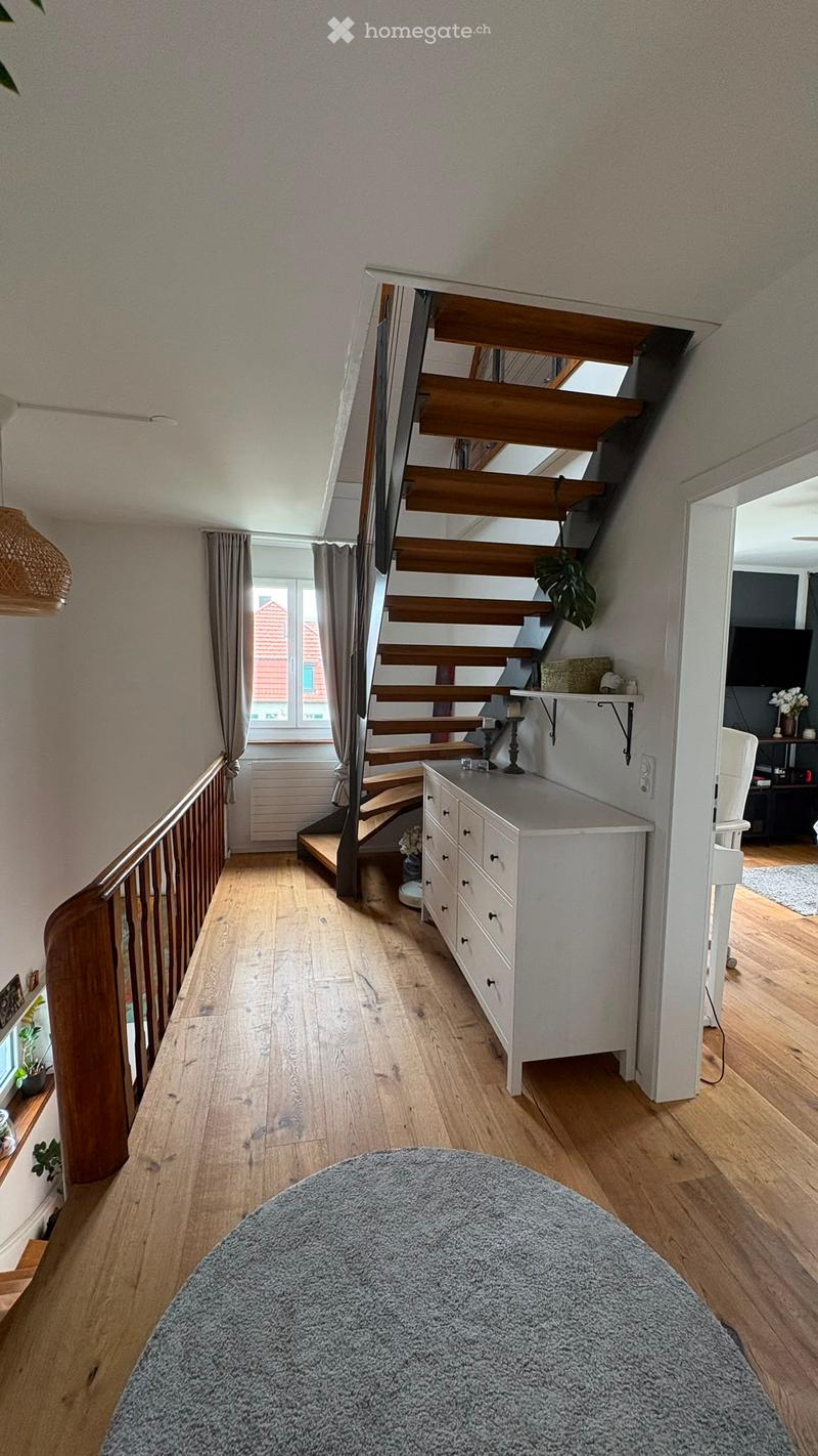 A modern hallway with wooden flooring, a staircase leading upwards, a white cabinet, a grey rug, and a hanging lamp.