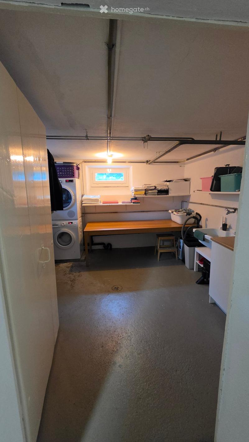 Laundry room with a washing machine, wooden table, small stool, sink, and shelves holding folded clothes and cleaning supplies.