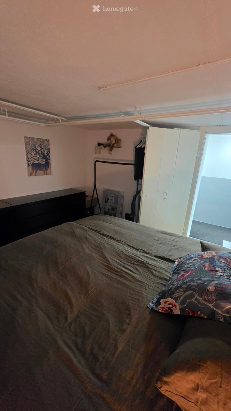 A small bedroom with a bed covered in a gray duvet and a colorful pillow, a wardrobe, a picture frame on the wall, and a small stool near the bed.
