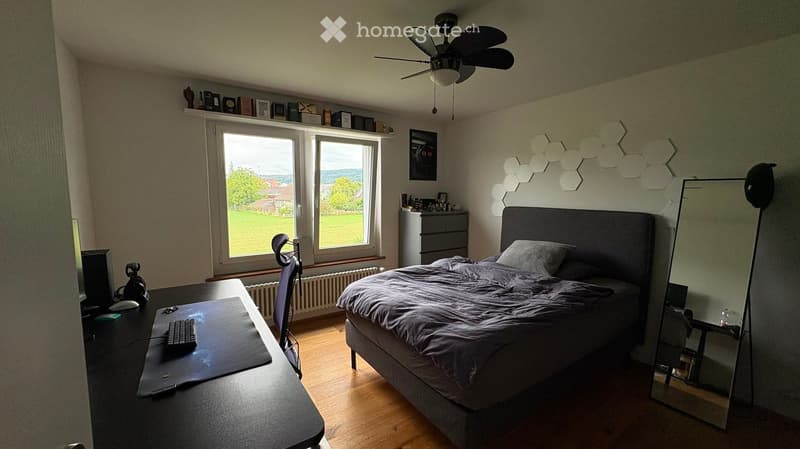 Well-lit bedroom with a large window offering a view of a green landscape. Features a bed with a dark headboard, a desk with a computer setup, a fan on the ceiling, and a cabinet with various items on top.