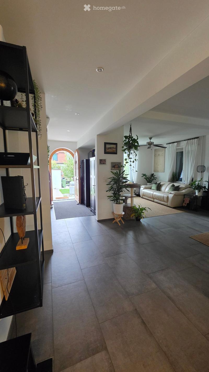 Spacious living room with dark tiled floor, white walls, and a high, slanted ceiling. Features a white sofa, cat tree, small table with plants, and a tall black shelving unit with books and decorative items.