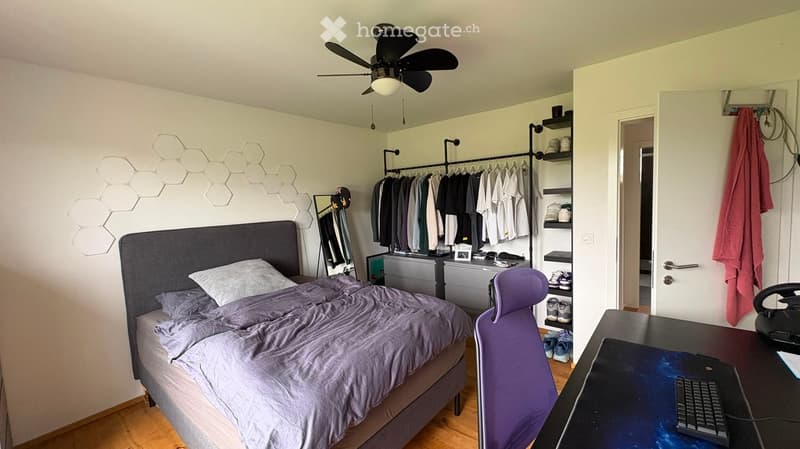 Modern bedroom with a grey bed, purple chair, desk with keyboard, and a clothing rack with hanging clothes. Features a hexagonal wall design and a ceiling fan with light.