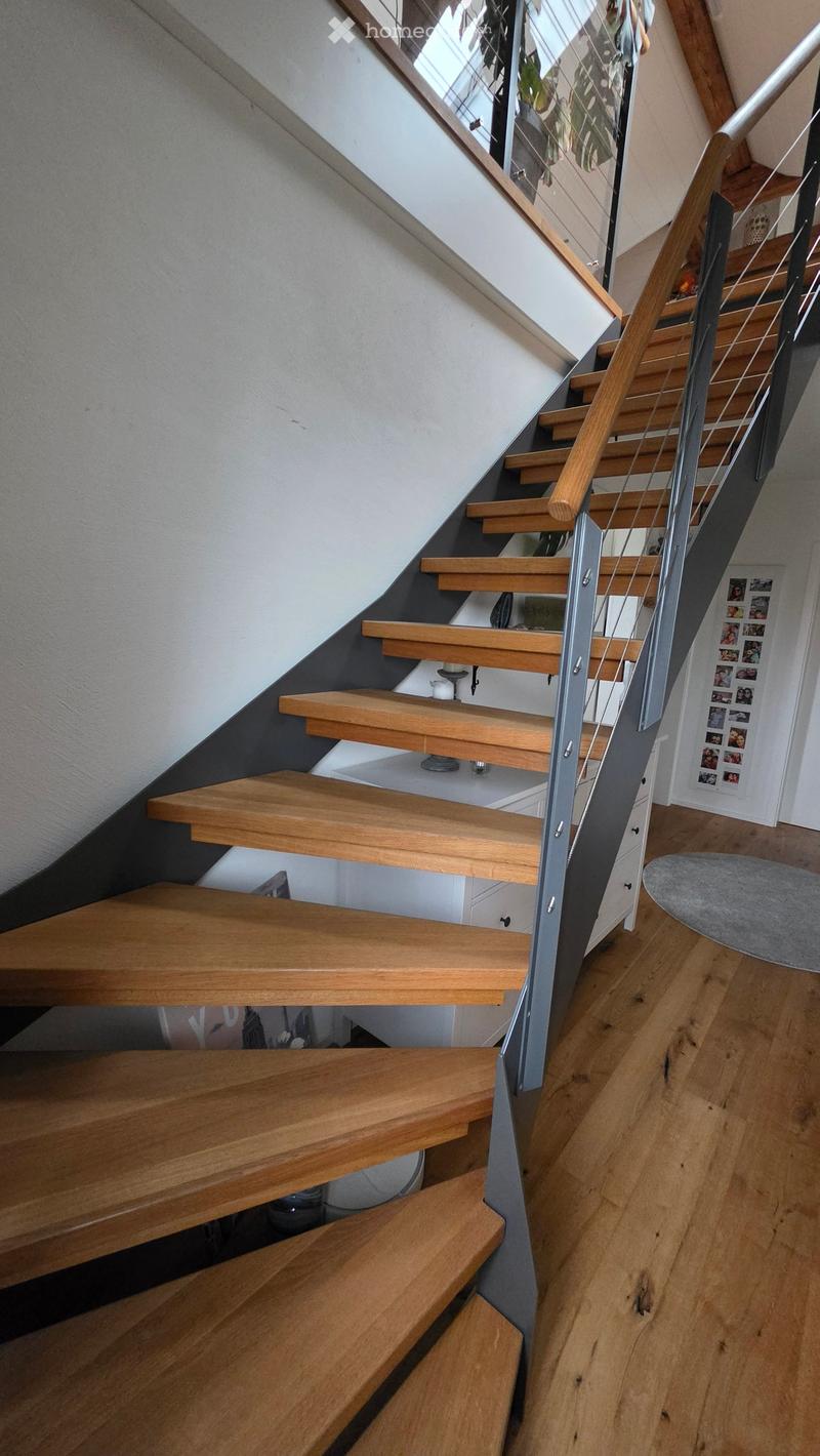 Modern wooden staircase with metal railing, leading to an upper level. The steps are light-colored wood, and the railing features a sleek metal design. The walls are painted in a neutral tone, and the floor is made of polished wood.