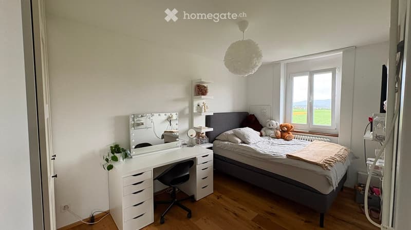Cozy bedroom with a large bed, a white desk with a chair, a vanity mirror, shelves with decorative items, and a view of a green landscape through the window.