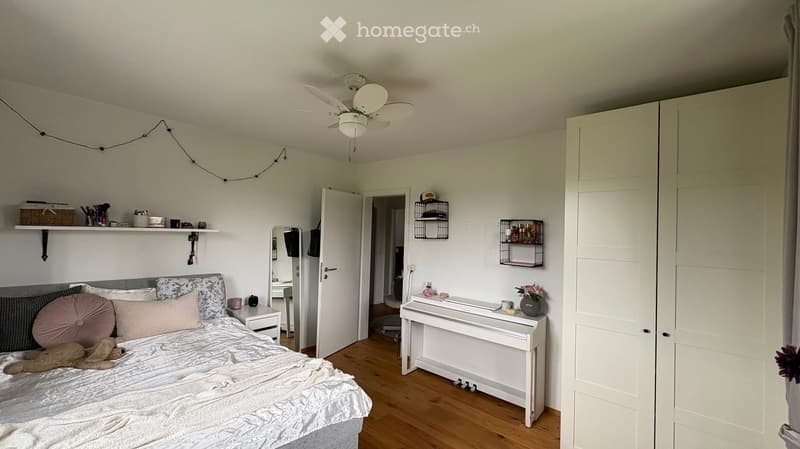 Cozy bedroom with a bed, piano, shelves, and a mirror. Features a wooden floor, white walls, and a ceiling fan.