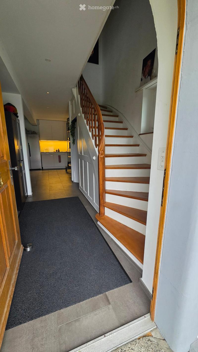 Staircase with wooden handrail and white steps, leading to a kitchen area with white cabinets and a yellow backsplash. The hallway has a dark carpet and a small rug at the base of the stairs.