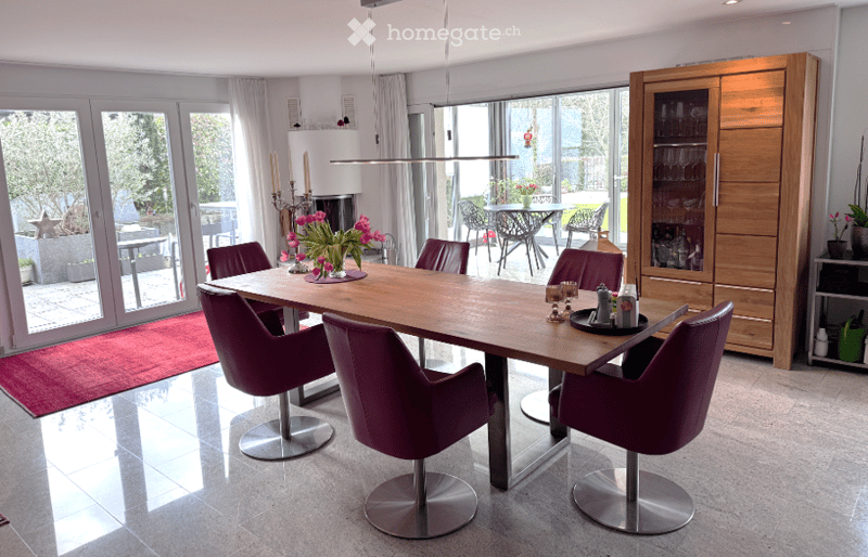Modern dining room with a wooden dining table surrounded by purple chairs, a wooden cabinet with glass doors, and a large window with a view of the garden.