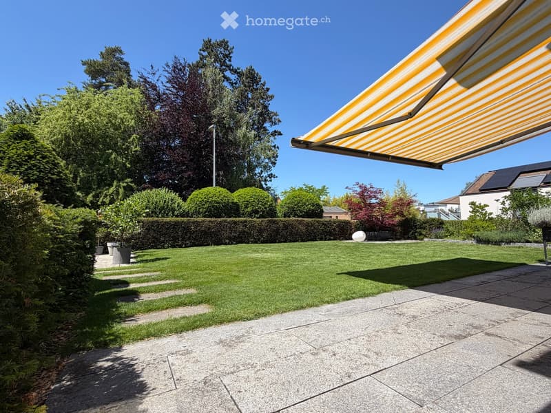 Well-maintained garden with a neatly trimmed lawn, a hedge border, potted plants, and a covered patio area with a striped awning.