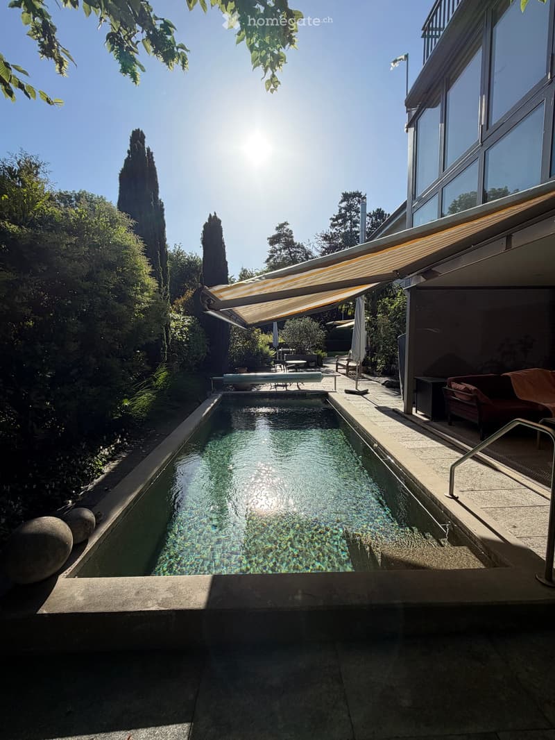 Outdoor area with a long, narrow swimming pool surrounded by greenery, a covered patio with seating, and a modern building in the background.