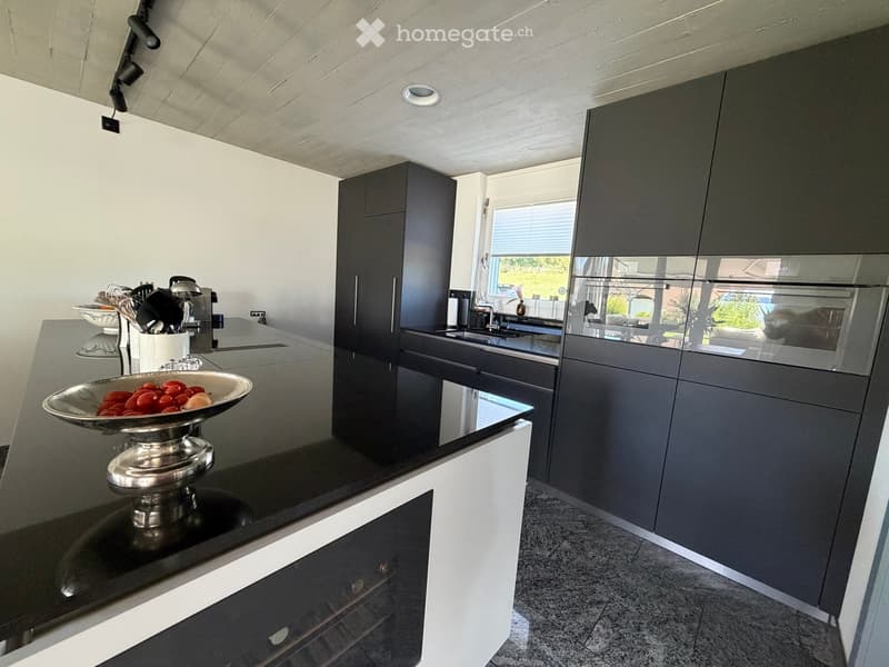 Modern kitchen with dark grey cabinets, black countertops, and stainless steel appliances. Features a bowl of fruit on the counter and a coffee maker.