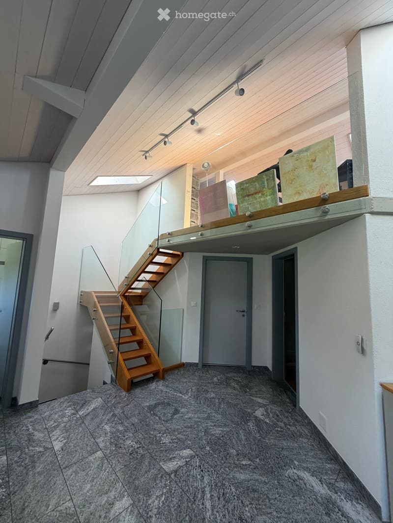 Modern hallway with a wooden staircase featuring glass railings, grey tiled floor, white walls, and recessed lighting.