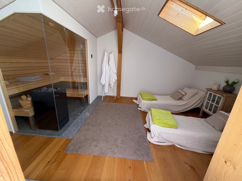 Cozy attic room with wooden sauna, two single beds, pillows, blankets, small table with vase, towels, and a basket.