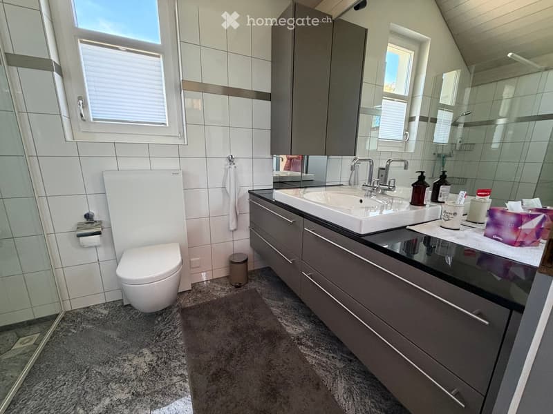Modern bathroom with white tiles, a double sink, a toilet, a large mirror, and a glass shower enclosure. The vanity has dark grey cabinets and a black countertop.