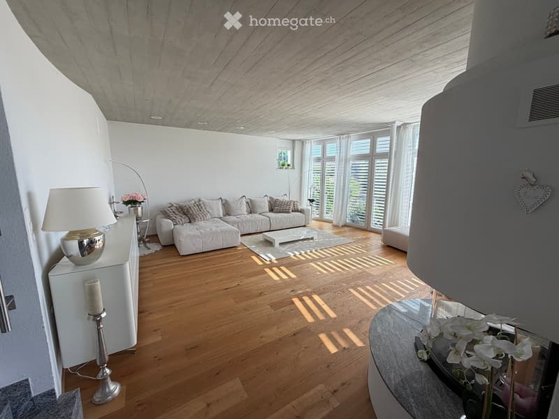 Modern living room with light wooden flooring, white walls, large windows, a white sectional sofa, coffee table, floor lamp, side table with vase, ottoman, and decorative candle holder.