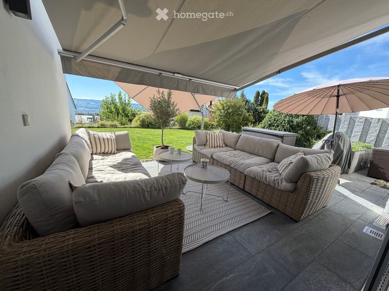 A modern outdoor patio with a large sectional sofa, wicker chairs, coffee tables, and an umbrella providing shade. The view includes a well-maintained lawn and distant mountains.