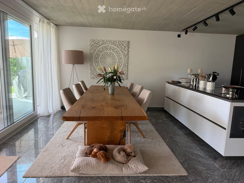 Modern dining area with a wooden dining table, six chairs, a vase with flowers, a kitchen counter with utensils, a floor lamp, and a cozy dog bed with a stuffed toy.