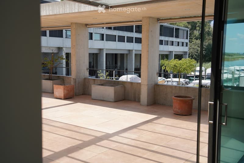 Modern outdoor patio with tiled flooring, large concrete planters, and a covered wooden pergola. The area overlooks a waterfront with boats and a modern building in the background.
