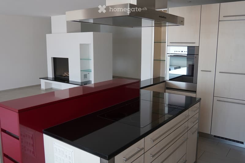 Modern kitchen with a red and black color scheme, featuring a central island, built-in stove, oven, range hood, and a fireplace in the background.