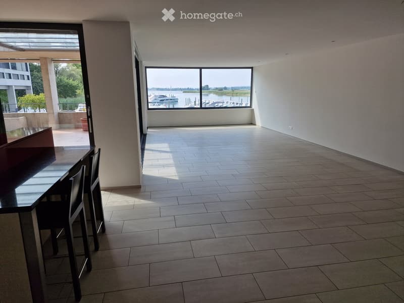 Bright, modern room with tiled flooring, large windows overlooking a waterfront, and a small dining setup with a table and chairs.