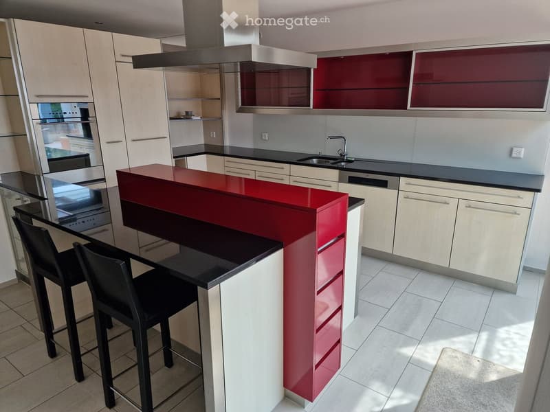 Modern kitchen with white and red cabinets, a central island with a red countertop, bar stools, and built-in appliances including an oven and sink.