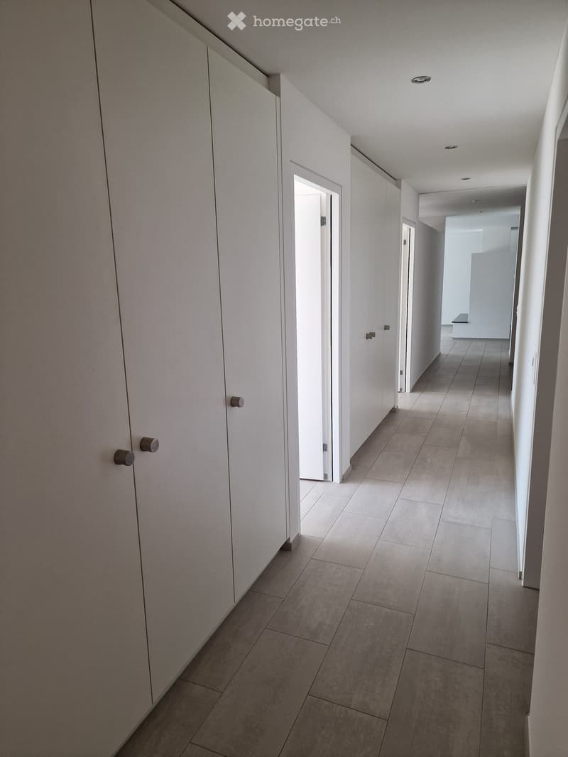 Modern hallway with white walls, tiled floor, and built-in storage units on one side. The hallway features recessed ceiling lighting and several doors leading to other rooms.