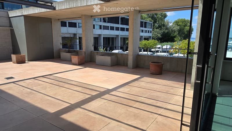 Modern outdoor patio with tiled flooring, concrete planters, and a covered wooden pergola. The area is open and spacious, with views of surrounding buildings and greenery.