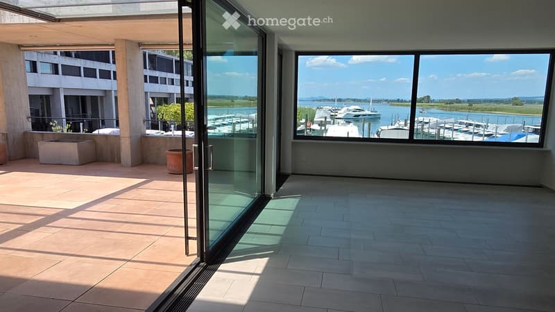 Spacious balcony with large glass doors, tiled flooring, and a view of a marina with multiple boats and a body of water.