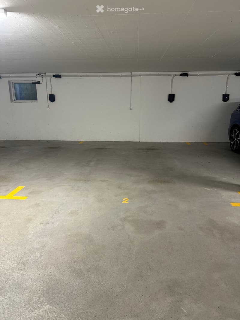 Empty underground parking space with concrete floor, white walls, and yellow parking markings. A small window and electrical outlets are visible on the wall.
