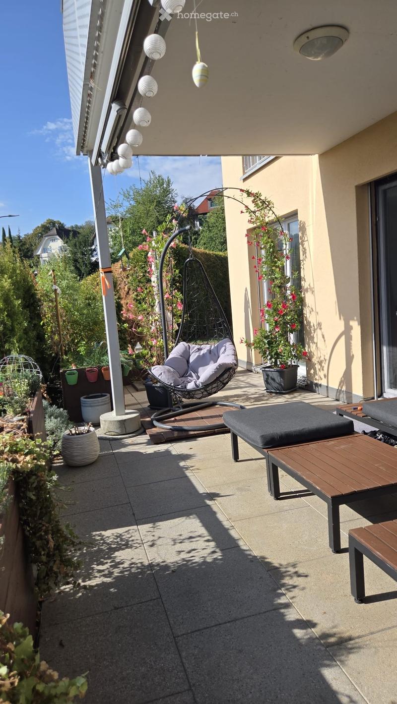 Balcony with a hanging egg chair, wooden table, wooden bench, and several potted plants.