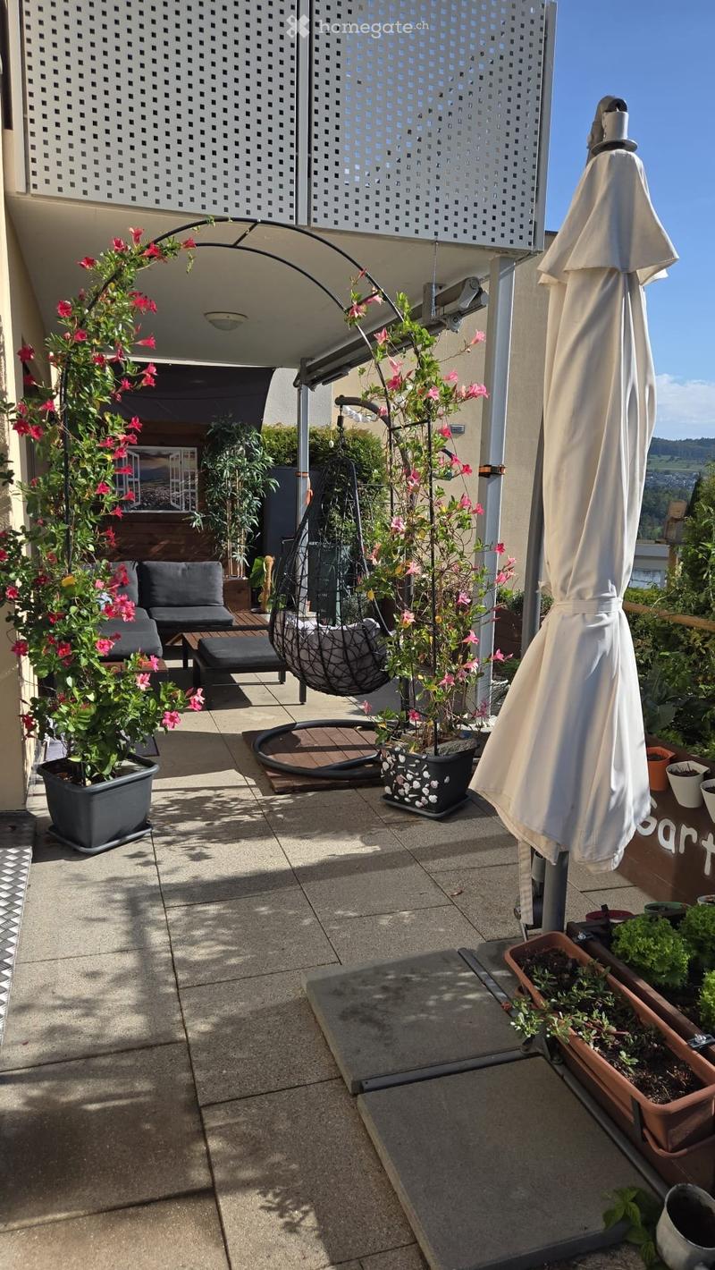 Balcony with potted plants, a hanging chair, a closed umbrella, and a small seating area with a sofa and table.