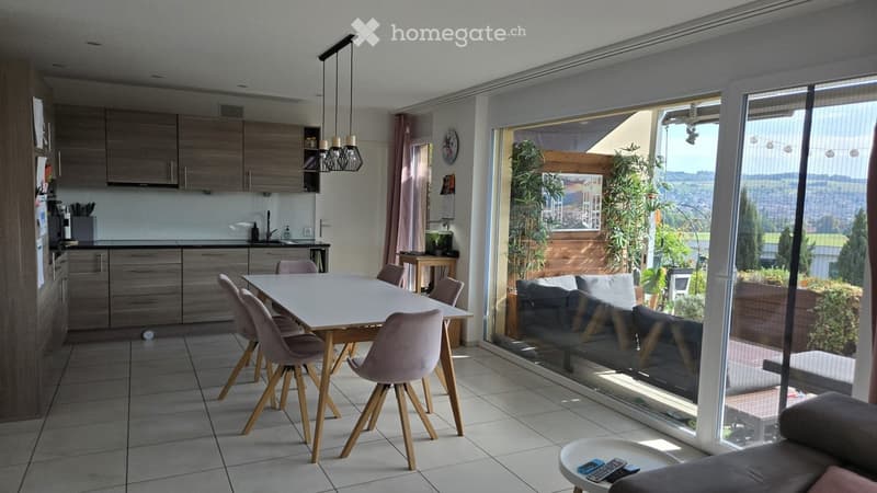 Bright, modern dining and living area with a white dining table, pink chairs, grey sofa, coffee table, and a fully equipped kitchen with wooden cabinets and stainless steel appliances. Large windows offer a scenic view of the landscape.