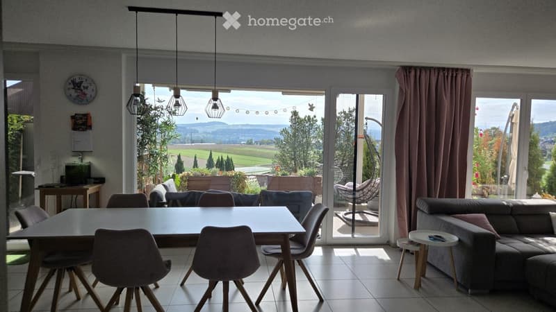 Bright living and dining area with large windows and a view of the countryside, featuring a sofa, dining table with chairs, and a small side table with a lamp.