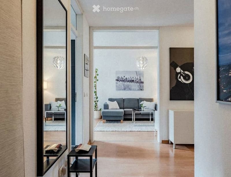 Modern living room with grey sofa, coffee table, side tables, potted plant, picture frames, and a mirror. The space features wooden flooring and large windows.