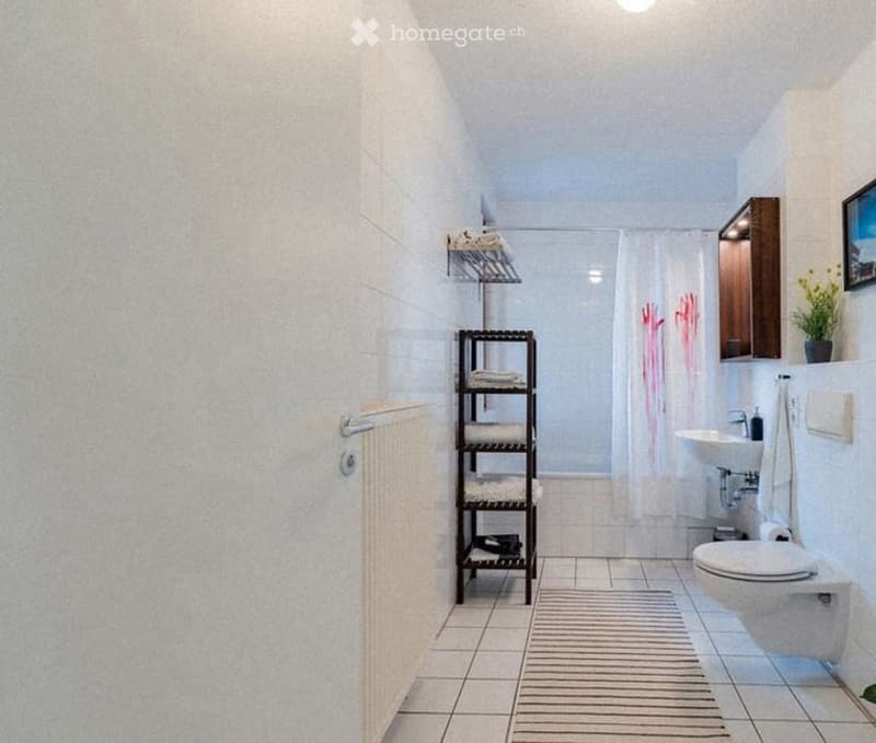 Modern bathroom with white tiled walls and floor, a shower area with a curtain, a sink with a cabinet, a toilet, a towel rack, and a small plant on the windowsill.