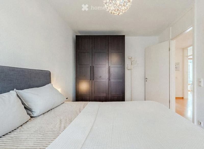Modern bedroom with a large bed, dark wooden wardrobe, and a coat rack near the door. The room features white walls and a decorative chandelier.