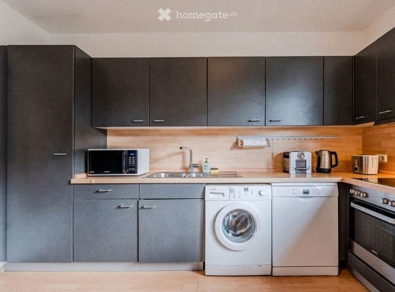 Modern kitchen with grey cabinets, white appliances including a microwave, washing machine, dishwasher, and oven, and a light wooden countertop.