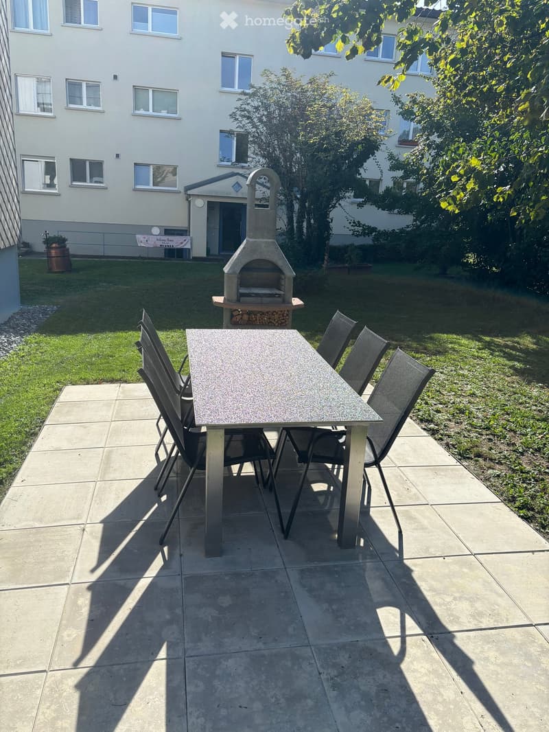 Outdoor patio with a stone pizza oven, a rectangular table, and four chairs, surrounded by a grassy lawn and trees.