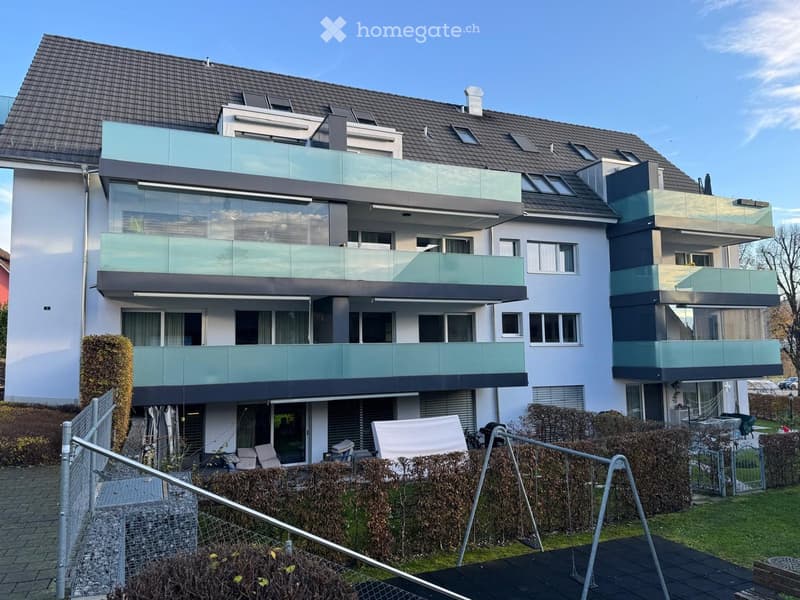 Modern two-story house with white and grey exterior, multiple large windows, and glass balconies. Features a fenced front yard with patio furniture and a small garden area.