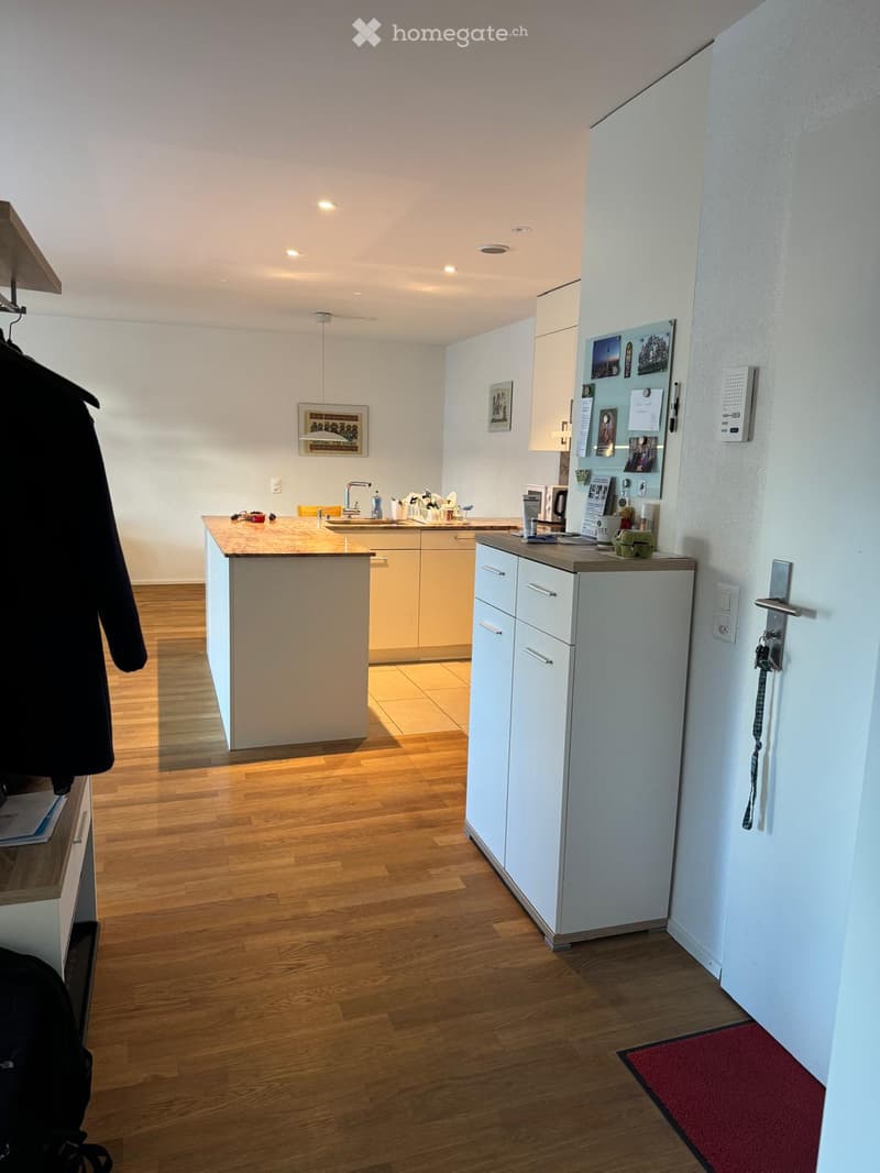 Modern kitchen with light-colored cabinets, a central island, and a refrigerator. The floor is wooden, and there are framed pictures on the wall.