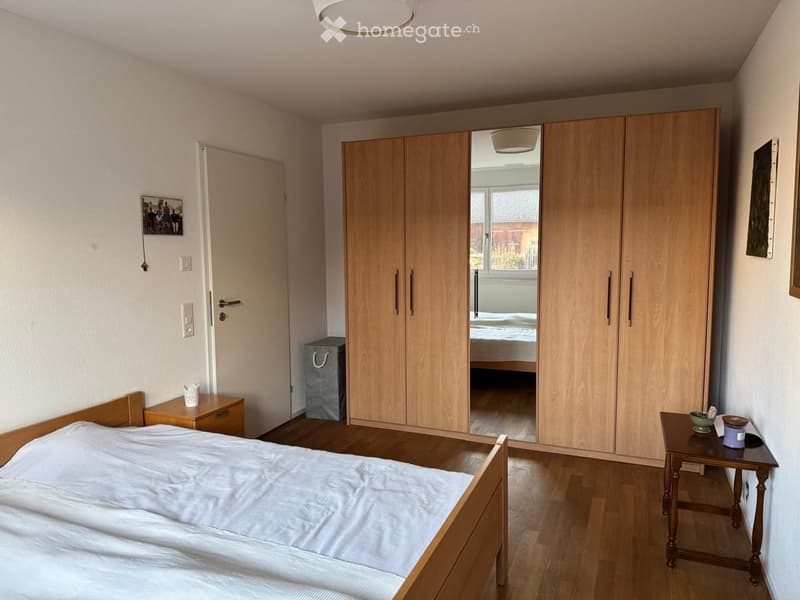 Well-lit bedroom with wooden bed, nightstand, wardrobe, small table with items, and a view into another room.