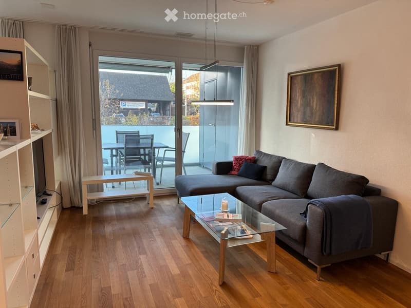 Modern living room with wooden flooring, grey sectional sofa, glass coffee table, white shelf unit, and a dining area visible through large glass doors.