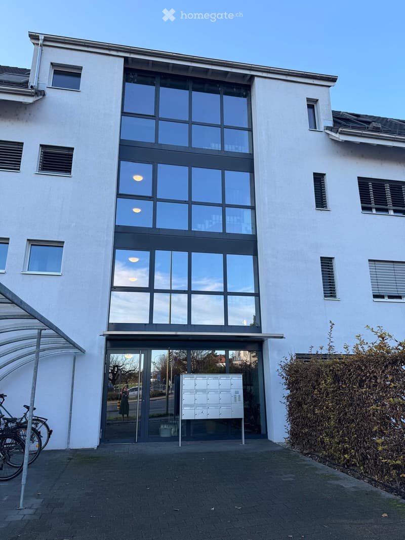 Modern two-story building with large glass windows and a central entrance, featuring a mailbox stand and bicycles parked nearby.