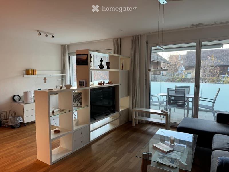 Modern living room with wooden flooring, large windows, a grey sofa, glass coffee table, shelves with decorative items, and a TV stand.