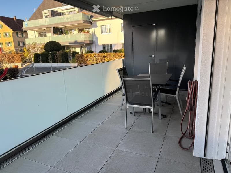 Balcony with tiled floor, glass railing, and outdoor furniture including chairs and a table.