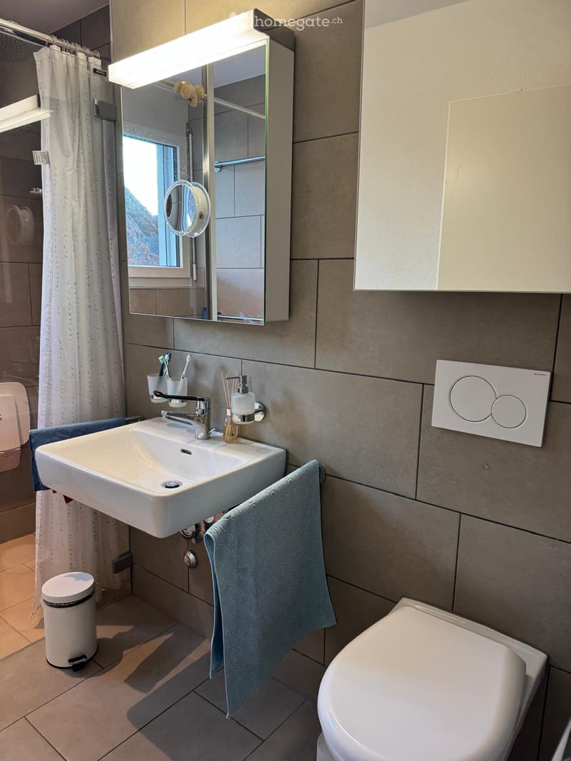 Modern bathroom with a white sink, mirror with built-in lighting, toilet, towel, and a small trash bin. The walls are tiled in a neutral color, and there is a window with a white curtain.