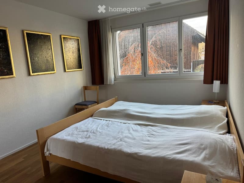 Cozy bedroom with a double bed, wooden nightstand, chair, and framed mirrors on the wall. Large windows with curtains let in natural light.